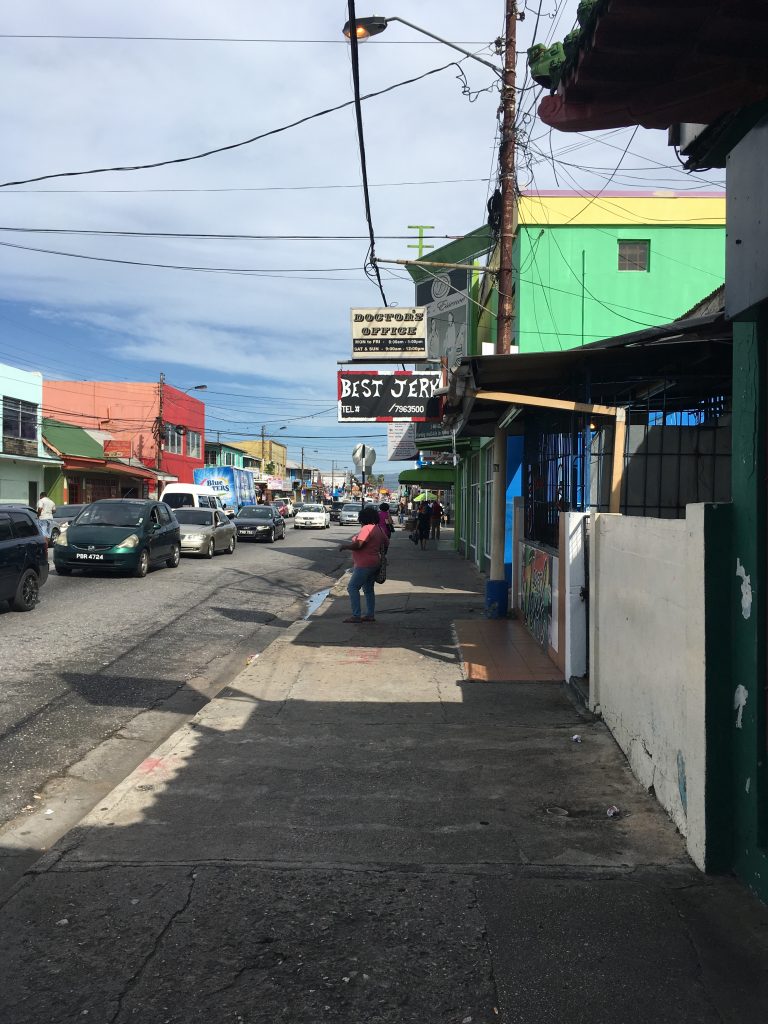 A colour image capturing the Eastern Main Road in St. James, Port of Spain during the day. There is a sign advertising the Best Jerk.